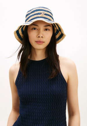 Ung kvinde med langt mørkt hår iført en blå og beige stribet bucket hat og en mørkeblå, ærmeløs strikketop.