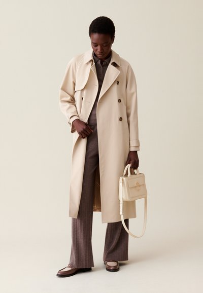 Cappotto beige con design doppiopetto, abbinato a pantaloni marroni a righe sottili e una borsa leggera. Texture liscia, accentuazioni hardware minime.