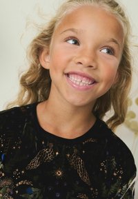 Melnā krekls ar zelta un zaļu ziedu izšuvumu, ar apaļu kakla izgriezumu un īsām piedurknēm, to valkā bērns ar viļņainiem gaišiem matiem.