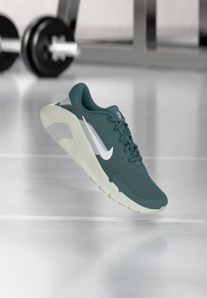 Turquoise Nike sportschoen met wit Swoosh-logo en lichte zool, zwevend in de lucht op een fitnessvloer met wazige gewichten op de achtergrond.