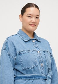 Denim shirt dress in light blue with a collared neckline, front buttons, chest pockets, and a tied waist belt. Soft, textured fabric.