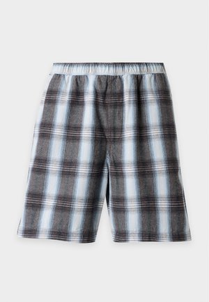 Mann steht mit den Händen in den Taschen, trägt ein blau-schwarz kariertes Hemd mit Knöpfen und passende karierte Shorts mit schwarzen Schuhen und weißen Socken.