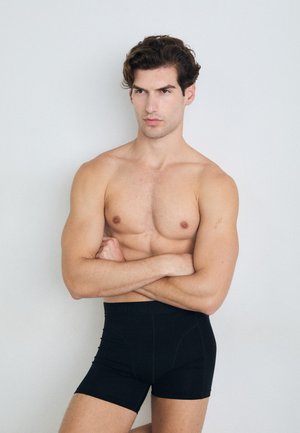Schwarz boxer Shorts aus glatter, dehnbarer Stoff, mit einem elastischen Bund mit Logo und einer enganliegenden Passform, getragen von einem hüllenlosen Modell mit verschränkten Armen.