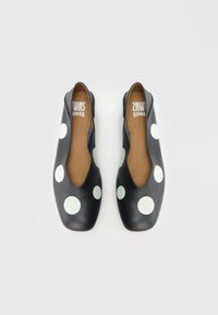 Zwarte platte schoenen met grote witte polka dots. Gladde, matte textuur, uitsnijdingen aan de zijkanten, en een afgeronde neus. Bruine leren binnenzool.