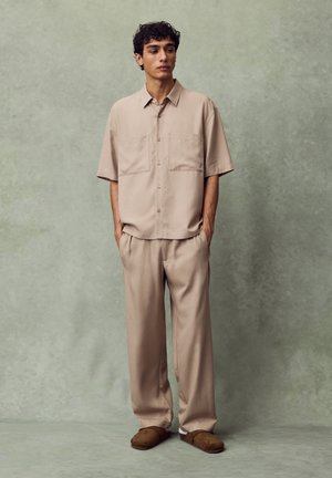 Mladi moški stoji z rokami v žepih, oblečen v bež srajco s kratkimi rokavi na gumbe, ujemajoče hlače in rjave natikače.
