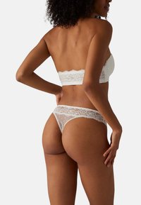 Bralette de encaje blanco y tanga a juego, con borde ondulado, lunares y paneles transparentes, mostrando un diseño delicado e intrincado.