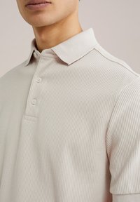 Polo krekls gaiši bež krāsā ar ribotu tekstūru, trīs pogas uz apakšžaketes un apkakles. Īsas piedurknes pabeidz dizainu.