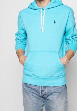 Homme portant un sweat à capuche bleu clair avec des cordons blancs et une poche avant, associé à un jean en denim bleu clair.