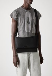 Borsa messenger in pelle nera con texture liscia, dotata di chiusura a patella e un discreto logo decorativo. Abbinata a una T-shirt grafica grigia e jeans chiari.