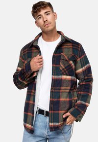 Camisa de franela en cuadros verdes, azul marino y naranja, con cuello abotonado, bolsillo en el pecho y forro interior suave. El modelo lleva una camiseta blanca.