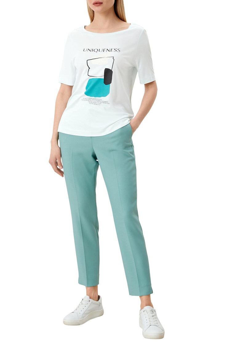 Witte grafische T-shirt met de tekst "UNIEKHEID", turquoise accenten, korte mouwen, gecombineerd met lichtblauwe broek en witte sneakers.