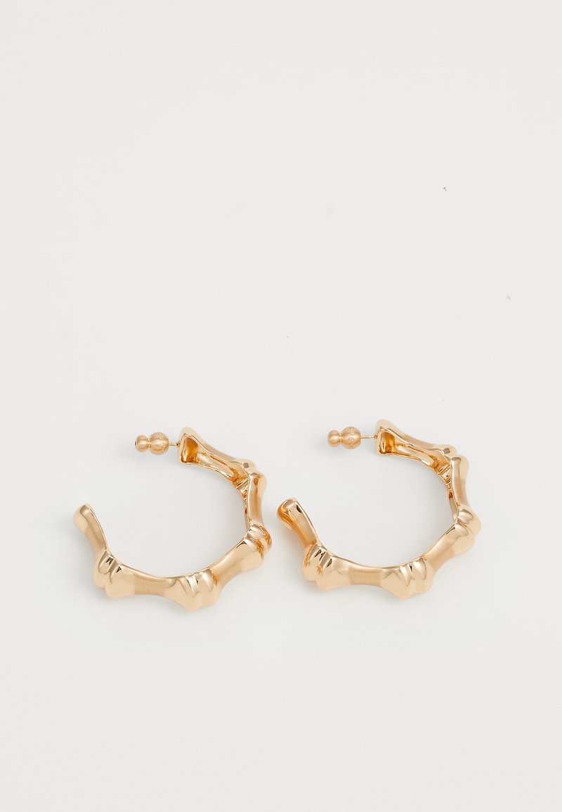 Χρυσά σκουλαρίκια κρίκοι με υφασμάτινη, κυματιστή σχεδίαση και λεία επιφάνεια. Διαθέτει κλείσιμο με κλιπ και μια μικρή διακοσμητική χάντρα.