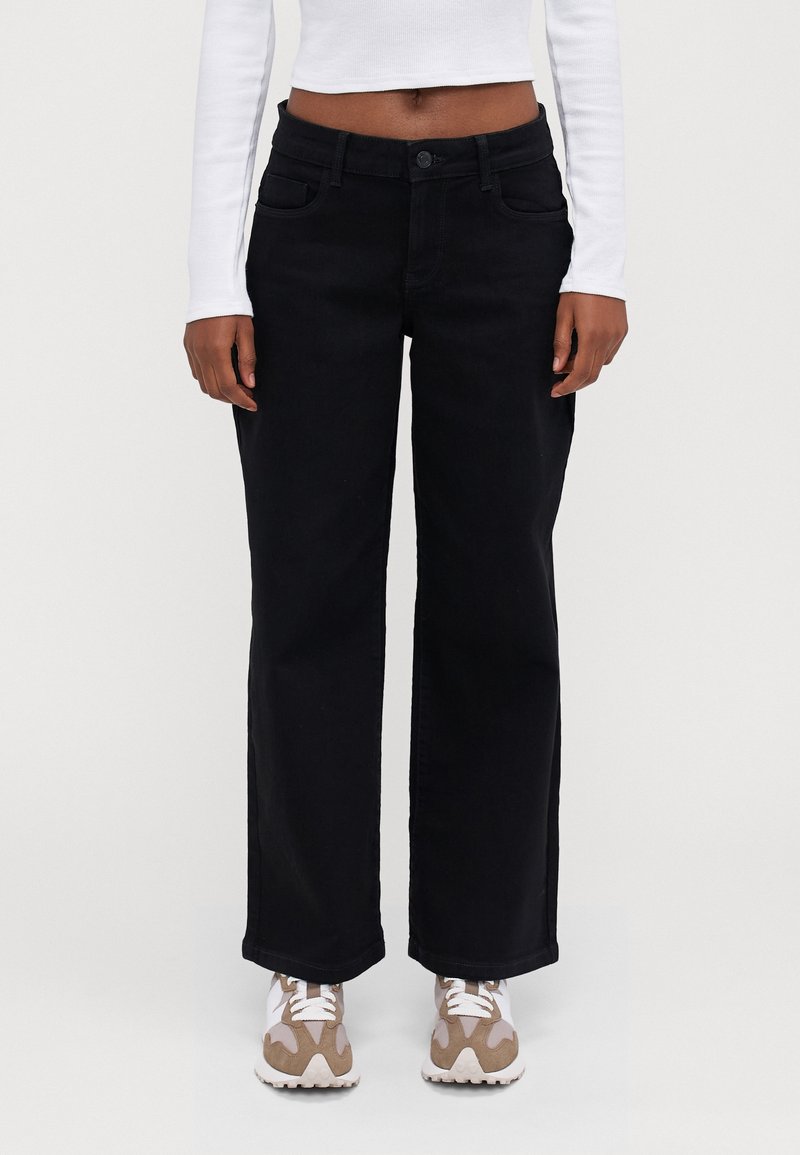 Czarne jeansy o szerokich nogawkach z zapięciem na guzik, przednimi kieszeniami i prostymi liniami. W połączeniu z białą bluzką z długim rękawem i brązowymi trampkami.