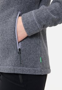 Giacca in pile grigia con tasca con zip, foderata in contrasto di grigio chiaro e etichetta verde del marchio sull'orlo. Presenta un tessuto testurizzato.