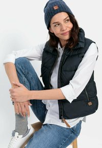 Gilet en duvet noir avec fermeture éclair, porté sur une chemise blanche. Associé à un jean bleu, des chaussettes grises et des baskets blanches. Bonnet en tricot bleu marine sur la tête.
