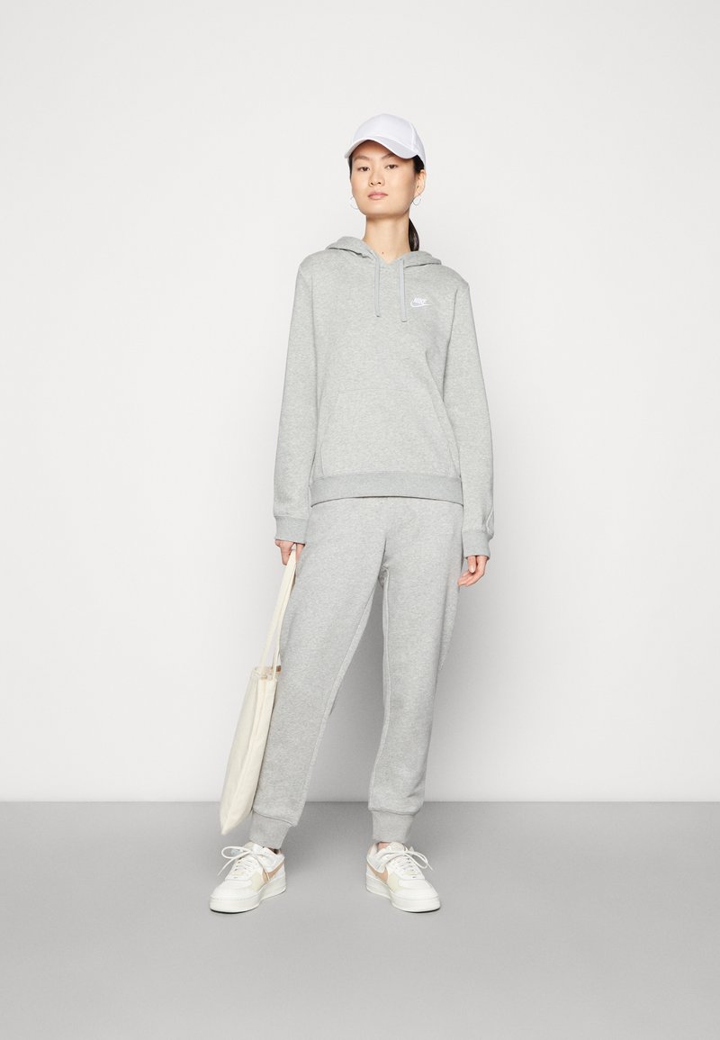 Graues Kapuzen-Sweatshirt und Jogginghose-Set mit einem Logo, aus weichem Material und mit gerippten Bündchen, kombiniert mit einer weißen Baseballkappe und einer Tragetasche.