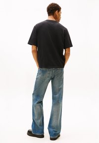 Camiseta negra de algodón con mangas cortas, combinada con vaqueros acampanados de color azul claro con lavado desgastado y bolsillos traseros. El calzado es oscuro y robusto.