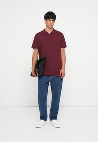 Burgunderfarget polo-skjorte med kortermet og krage, brukt med blå jeans og hvite sneakers. Svart veske i hånden, enkel design og passform.