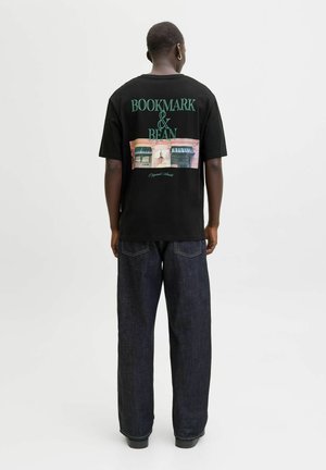 Férfi fekete pólóban, amelyen a "BOOKMARK & BEAN" felirat és egy épület grafika látható a hátán, sötét, széles szárú farmernadrágban, fehér háttér előtt áll.