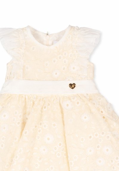 Vestido de bebé color crema con patrón de encaje floral, mangas cortas con volantes y una cinturilla blanca con un pequeño emblema de corazón dorado.