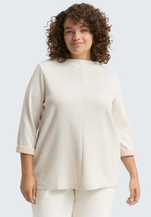 Vrouw met krullend haar, gekleed in een crèmekleurig ribbloes met lange mouwen en opgerolde manchetten, evenals bijpassende crèmekleurige broek, staande tegen een lichtgrijze achtergrond.