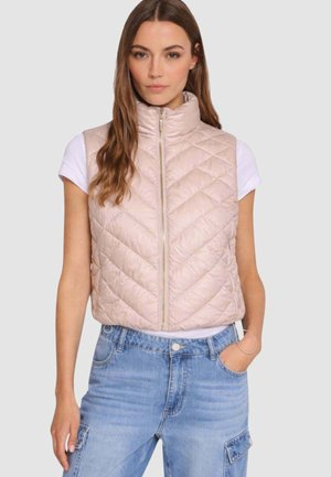 Donna che indossa un gilet trapuntato beige chiaro senza maniche sopra una maglietta bianca e jeans cargo a vita alta azzurri con la mano in tasca.