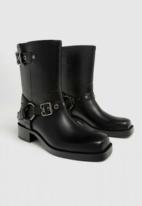 Botas negras de cuero hasta el tobillo con un tacón grueso, detalles de hebilla y acentos de hardware circular; textura suave y diseño de caña corta.
