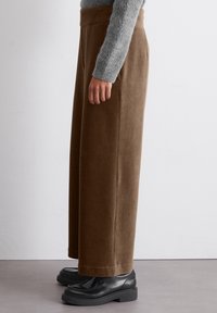 Person, der hoch taillierte, weit geschnittene braune Cordhosen, einen grauen Strickpullover und schwarze Plateauschuhe trägt, steht seitlich auf grauem Boden.