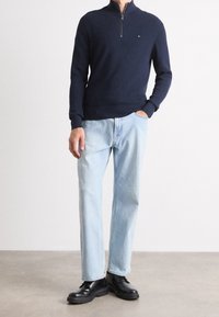 Marinblå stickad tröja med fjärdedels dragkedja, kombinerad med ljusblå raka jeans och svarta snörskor. Enkel, funktionell design.