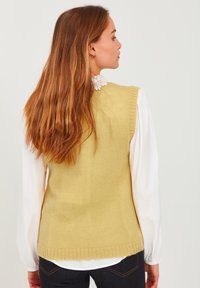 Knit sleeveless beige sweater with ribbed edges, worn over a white long-sleeve shirt. Features a light texture and a relaxed fit.
