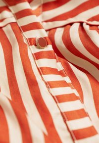 Gestreepte blouse met rode en crèmekleurige tinten, zachte stof, knoopdetails en zichtbare stiksels langs de knoopsluiting.