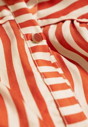 Gestreifte Bluse in Rot- und Cremetönen, glattes Material, Knopfdetail und sichtbare Nähte entlang der Knopfleiste.