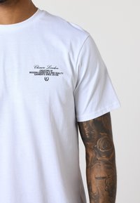 Weißes Baumwoll-T-Shirt mit einem kleinen schwarzen Logo-Text auf der linken Brust. Kurze Ärmel und eine lockere Passform mit einer glatten Textur.