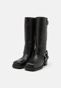 Bottes noires en cuir montant jusqu'au genou avec un bout arrondi, une fermeture éclair sur le côté, et une bride avec des éléments décoratifs en argent. Surface texturée et talon robuste.