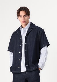 Mørk denim kortermet jakke med slagkrage og knaplukking, lagd over en lyse stripet langermet skjorte med hvite knapper.