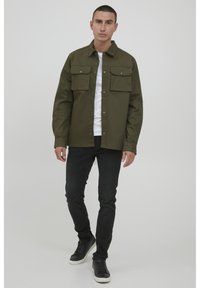 Chaqueta de botón verde oliva con dos bolsillos en el pecho, llevada sobre una camisa blanca. Combinada con jeans negros y zapatillas negras.