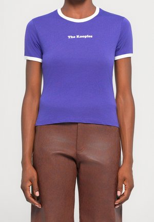 Vrouw draagt een aansluitend paars T-shirt met witte bies en de tekst "The Kooples", gecombineerd met hooggetailleerde bruine leren broek.