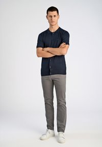 Marinblå kortärmad skjorta med knappar, grå slim-fit byxor och vita sneakers. Modellen står med armarna i kors mot en ljus bakgrund.
