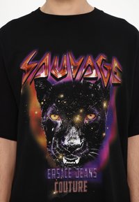 Juodas medvilninis marškinėlis, kuriame vaizduojama spalvinga panteros veido grafika, su geltonomis akimis ir kosmoso tema fono. Tekstas skelbia "SAUVAGE" ir "VERSACE JEANS COUTURE."