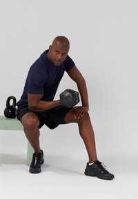 Dumbbell nera tenuta da una persona in una maglietta blu navy a maniche corte e pantaloni corti neri, seduta su una panchina verde pallido, con un kettlebell nero vicino.
