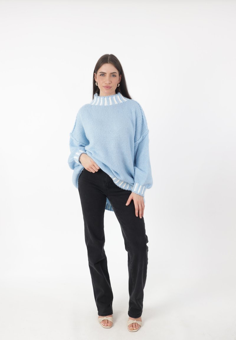 Lichtblauwe gebreide trui met een hoge hals en witte gestreepte details, gecombineerd met zwarte rechte jeans en beige sandalen.