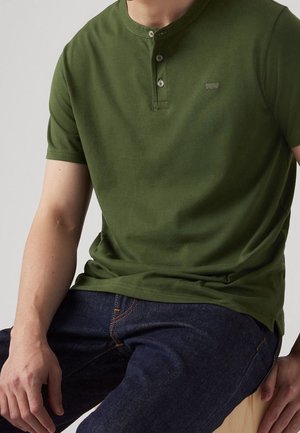 Man zit en draagt een olijfgroen henley-shirt met korte mouwen en drie knopen, gecombineerd met donkerblauwe spijkerbroek.