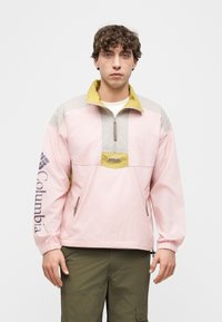 Νέος άνδρας με ροζ και μπεζ μπουφάν Columbia με λογότυπο στο μανίκι, συνδυασμένο με χακί παντελόνι cargο, στέκεται μπροστά σε απλό φόντο.