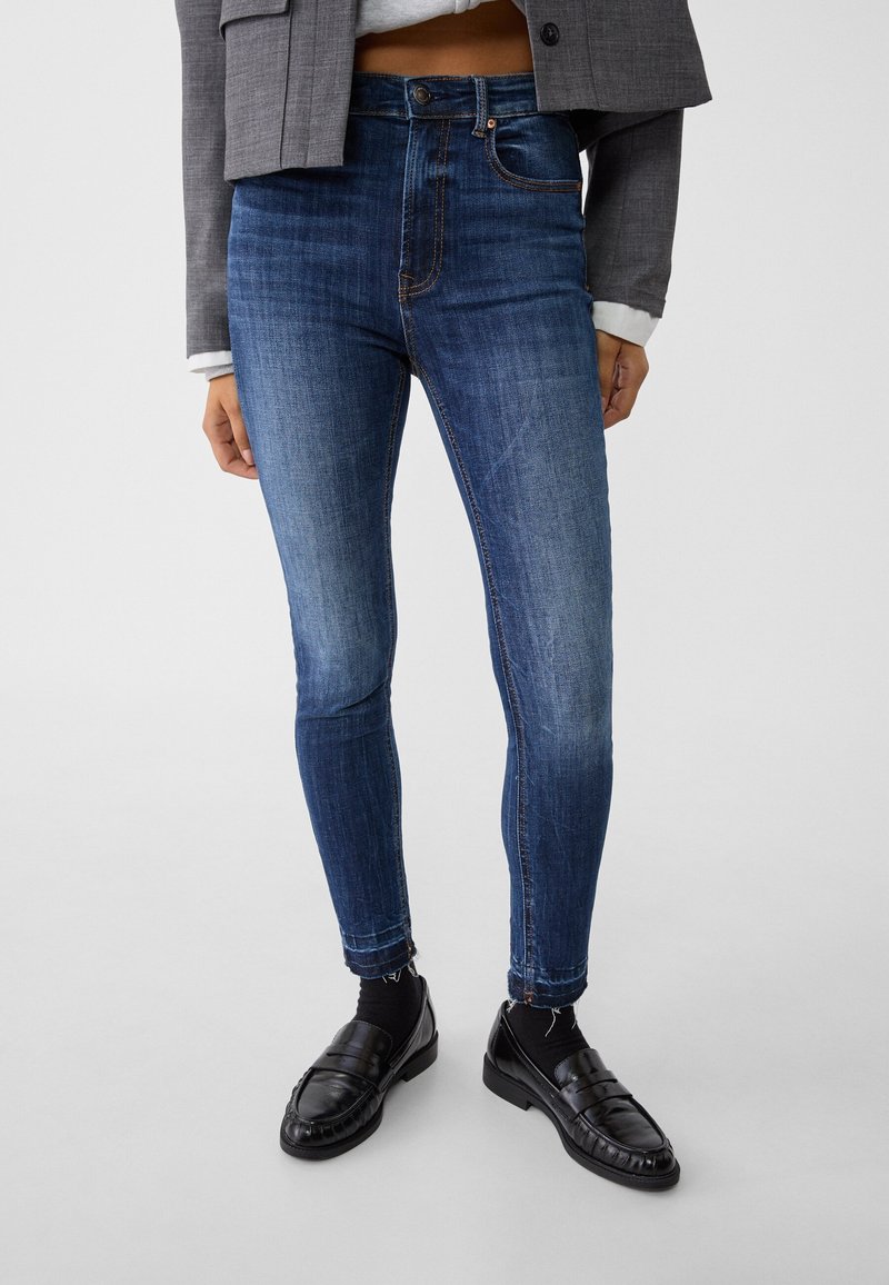 Jean skinny en jean bleu foncé avec des détails décolorés. Design taille haute, style à cinq poches, avec une fermeture éclair et des coutures contrastées.