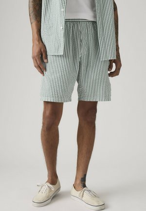 Mand iført grønne og hvide shorts med lodrette striber og en matchende skjorte over en hvid tanktop med hvide lærredssneakers.