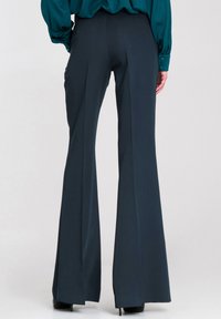 Kobieta w ciemnych, szerokich spodniach z wysokim stanem i turkusowej bluzce z długim rękawem, stojąca na jednolitym jasnym tle.