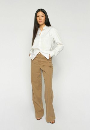 Pikajuustega naine, kes kannab valget nööbiga särk ja pruune laia säärega pükse, seisab käed taskutes ühevärvilise tausta ees.