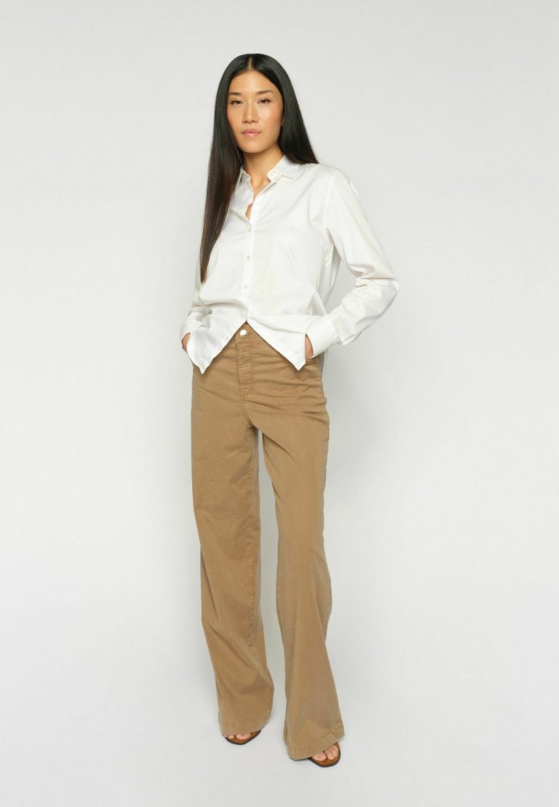 Hosszú fekete hajú nő fehér gombos ingben és barna, széles szárú nadrágban, zsebre tett kézzel, egyszínű háttér előtt áll.