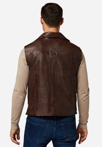 Braungefärbte Lederweste mit glatter Textur, spitzem Kragen und sauberer Naht. Kombiniert mit einem beigen Langarmhemd und blauen Jeans.