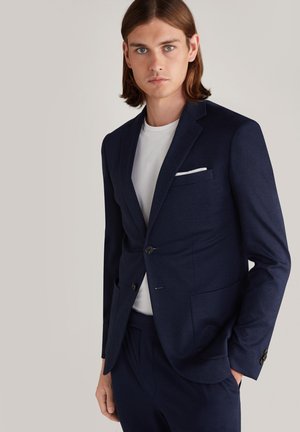 Navy blue tailored blazer with two front flap pockets, a single button, and a white pocket square, paired with a white crew-neck T-shirt.
